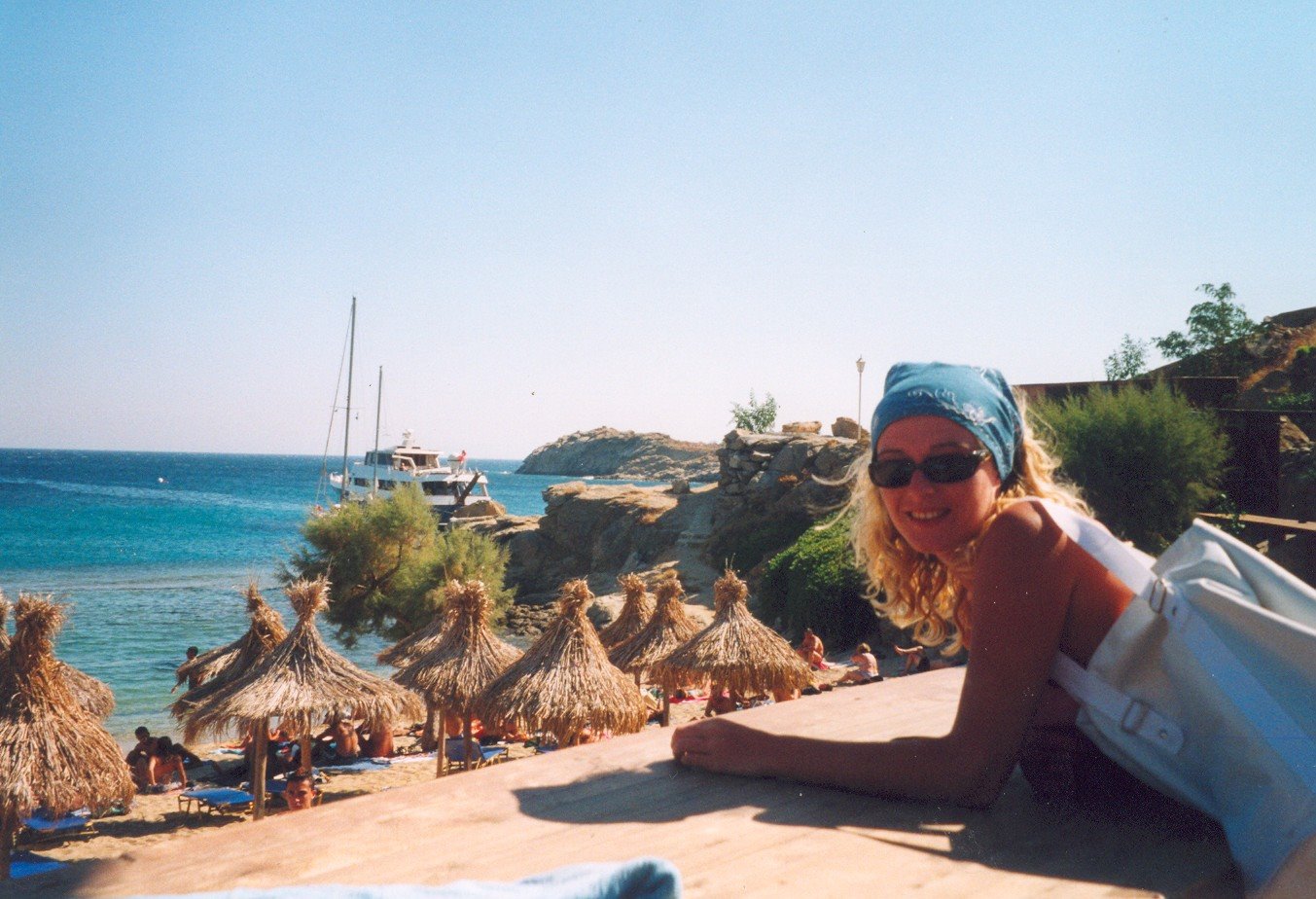 Ragazza con bandana e occhiali si sporge su terrazza di legno sopra spiaggia con ombrelloni di paglia