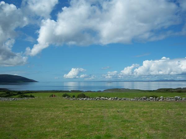 Dry stone wall across green pasture by calm sea under white clouds