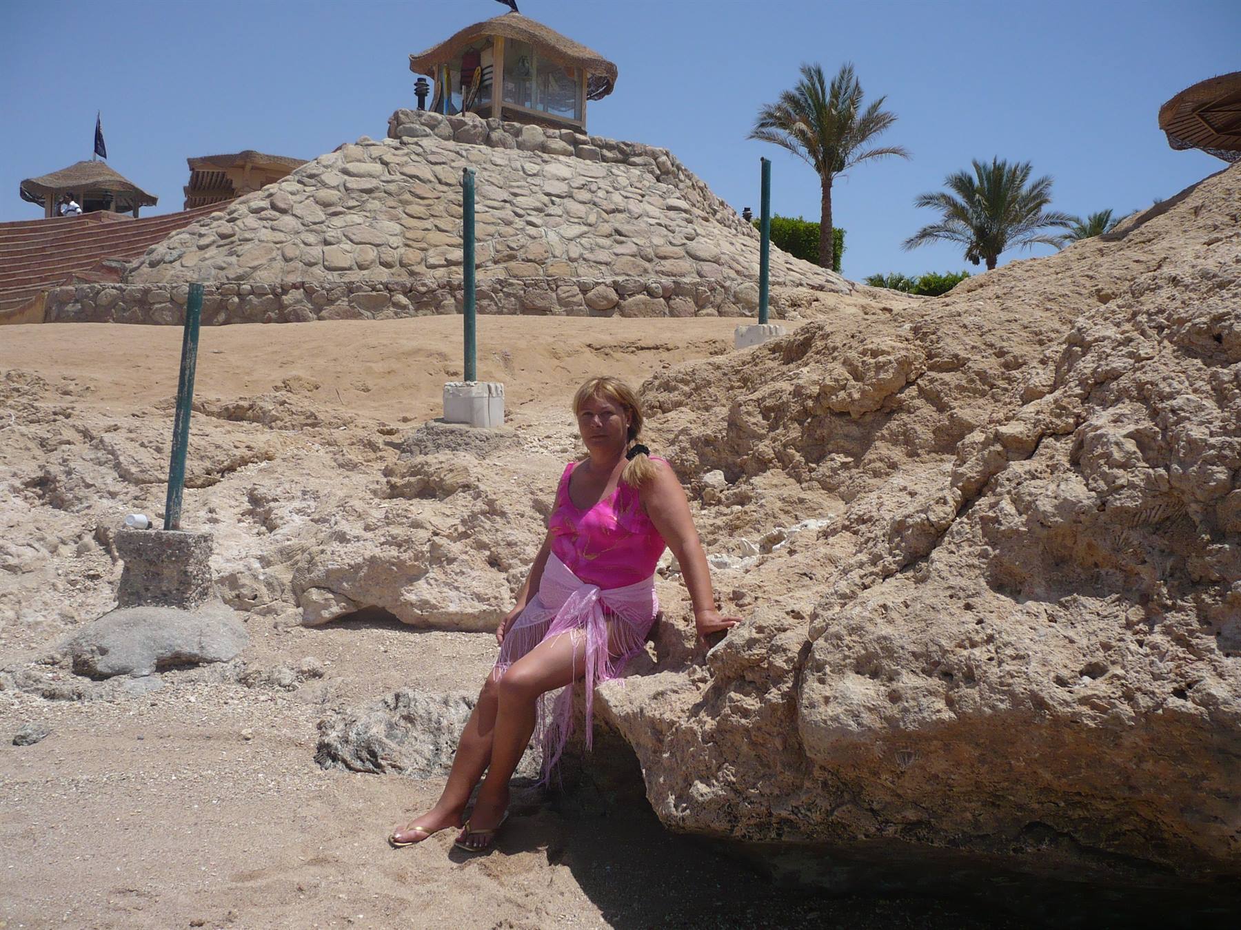 Woman in pink top sitting on rocky reef, palm trees and thatched kiosk behind