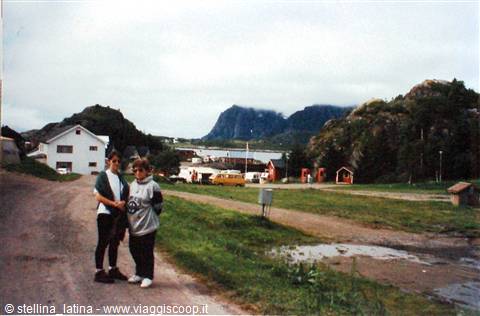 Two travelers on the gravel path among red rorbu cabins, Lofoten peaks behind