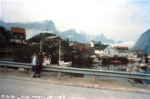 People by the road facing the harbor with boats, houses and misty mountains