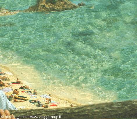 Beachgoers on sand beside rippling turquoise sea and rocky cliffs