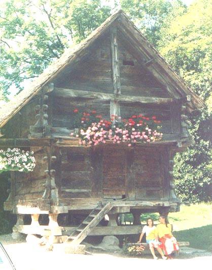 Two women sitting by an old wooden granary on stone stilts, flowers on the balcony