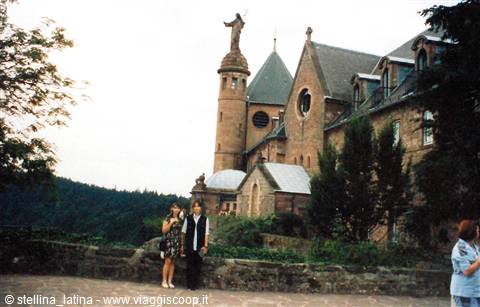 Tourists in front of the stone abbey with tower and statue, among trees