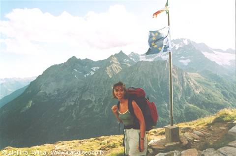 hiker with backpack beside Italian and EU flags on grassy summit, rocky mountains behind