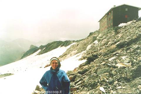 Girl in blue jacket on snow and rocks below a mountain hut