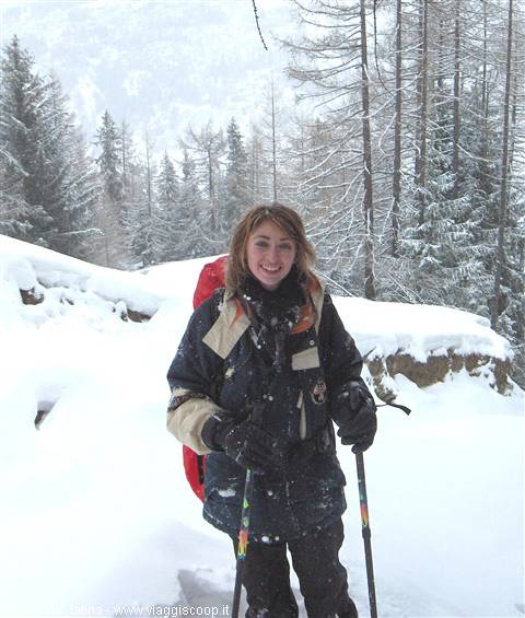 Smiling woman with red backpack and trekking poles walks through snowy firs