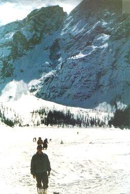 Hikers walk across snow in front of snowy rocky mountains and woods