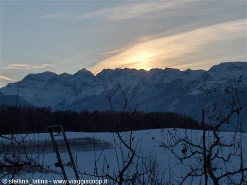 Sunrise behind snowy peaks, white field and bare branches in the foreground