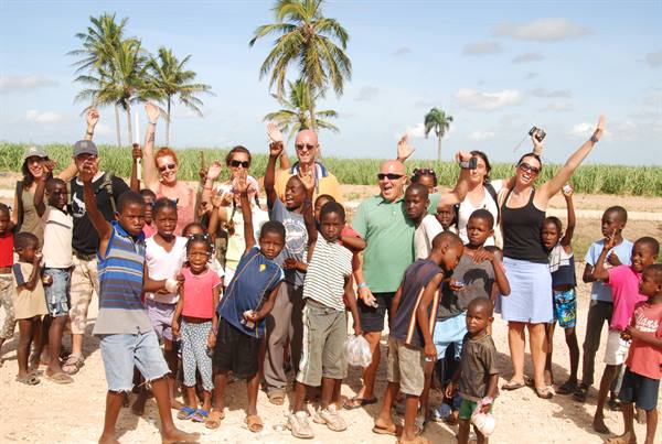 Group of tourists and local children pose arms raised among palms and fields