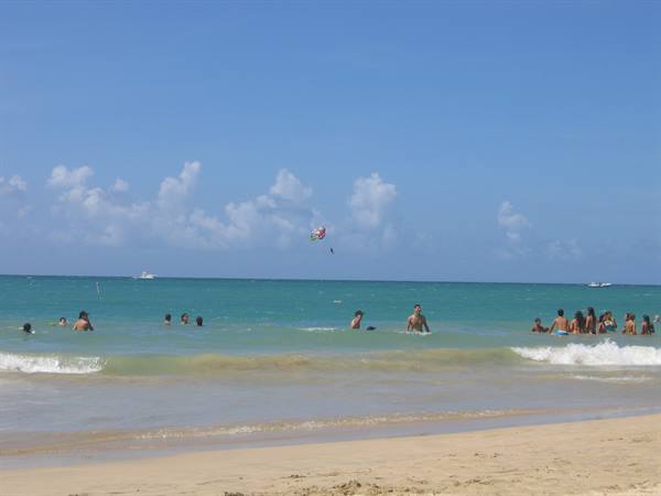 Swimmers in turquoise water, parasail flying above, light sand under blue sky
