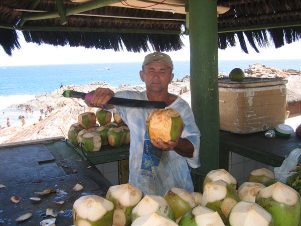 Man with machete cutting a coconut at a thatched beach kiosk