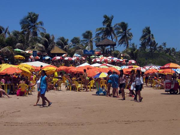 Crowded sunny beach with orange and yellow umbrellas, palm trees and beachgoers
