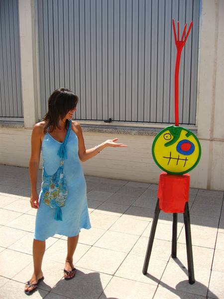 Woman in blue dress gestures toward a colorful Miró-style sculpture on tiled pavement