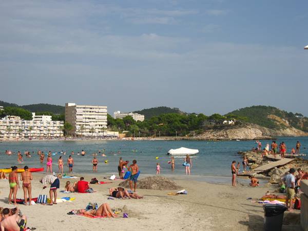 Tourists sunbathe on sandy beach, swimmers in calm water, hotels behind