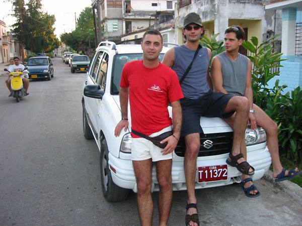 Three friends leaning on a white Suzuki car with Cuban plate, residential street