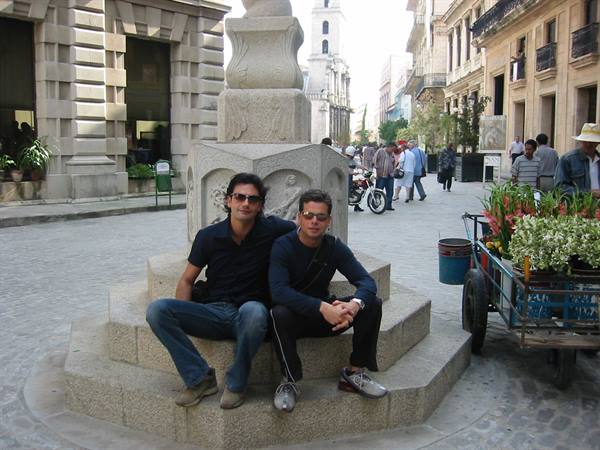 Two young men sitting on a stone fountain in a square with a flower cart