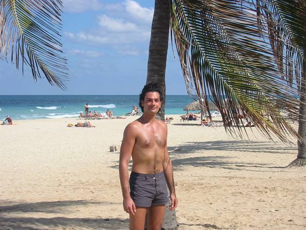 Man in gray swim shorts standing under a palm tree, white sand beach and turquoise sea