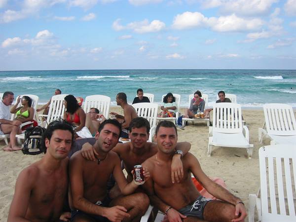 Four friends on a sandy beach with white loungers, one shows a can