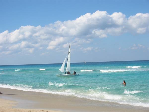 White sailboat on turquoise sea, swimmers and pale sand under clouds