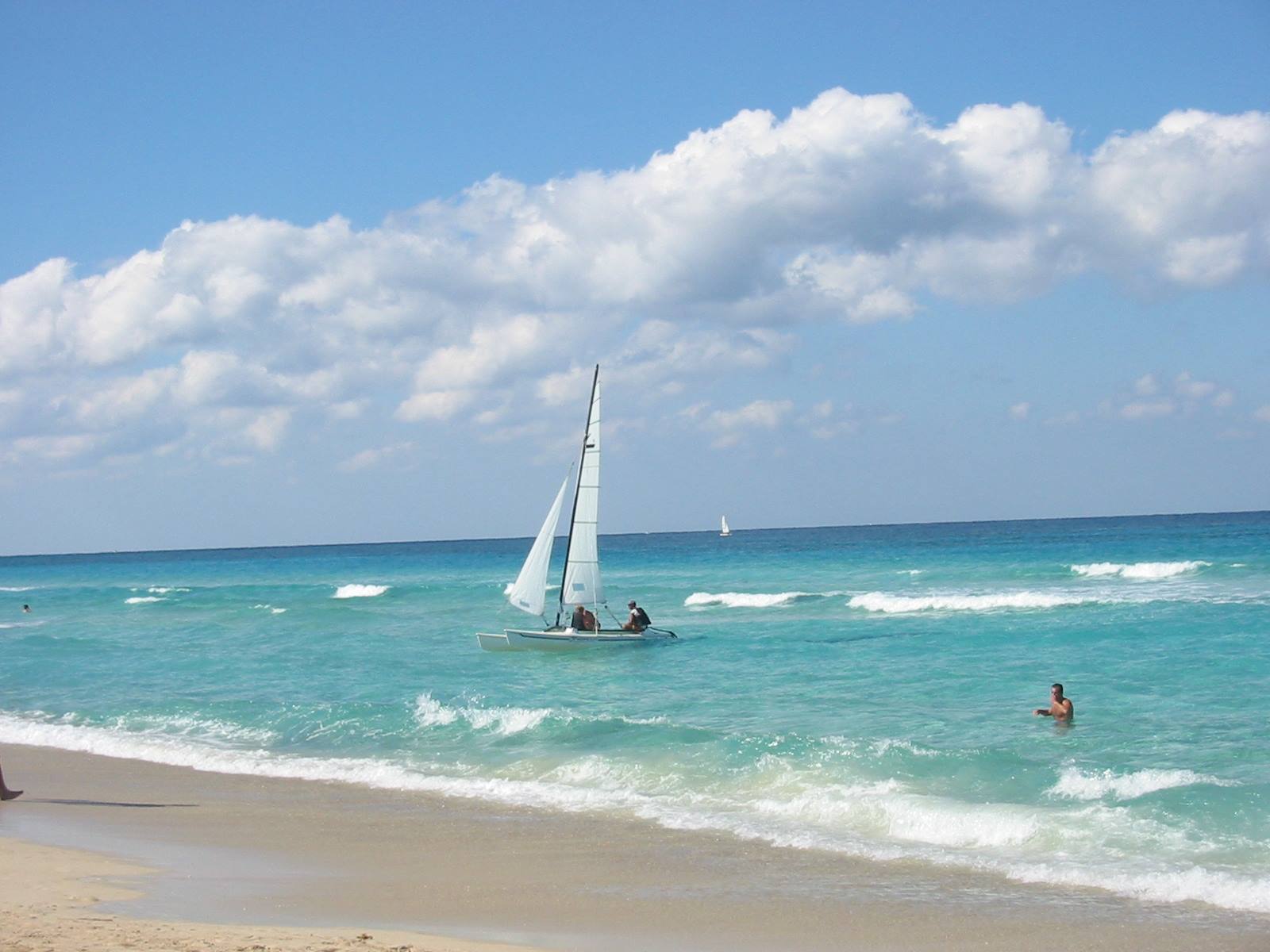 White sailboat on turquoise sea, swimmers and pale sand under clouds
