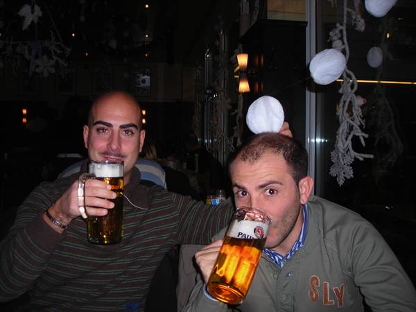 Two men toast with large Paulaner beer mugs in a bar decorated with white ornaments