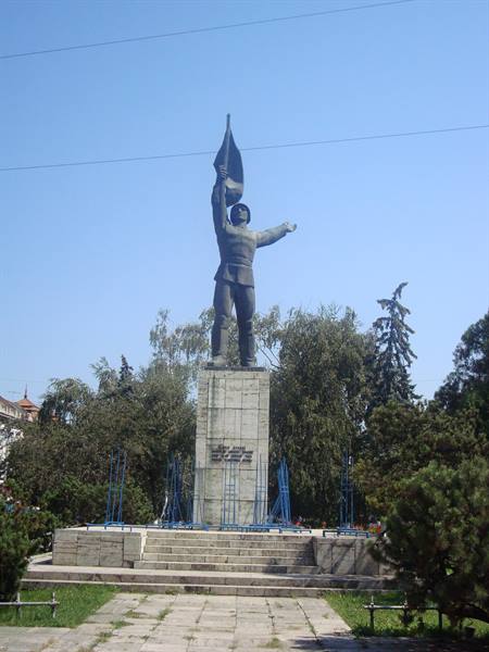 Statue of man raising a flag on stone pedestal, trees and blue sky