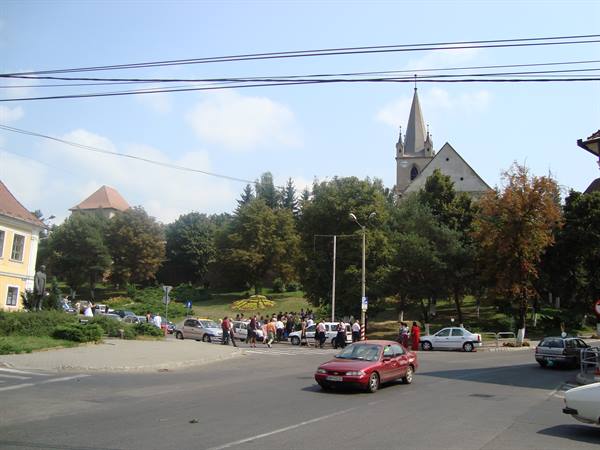 Cars and people cross a road in front of Sighișoara's Saint Michael church
