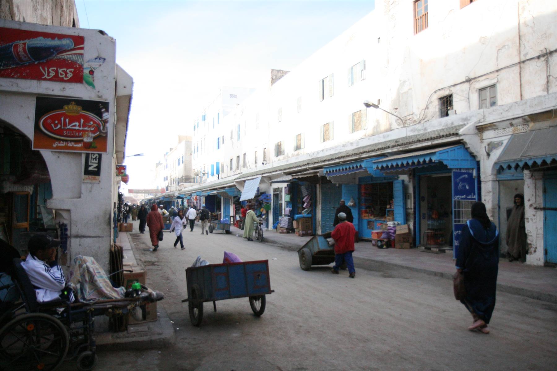 Medina street with market stalls, blue cart and passersby among whitewashed walls, Essaouira