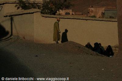 Man in green djellaba beside an earthen wall, women sitting in the shade
