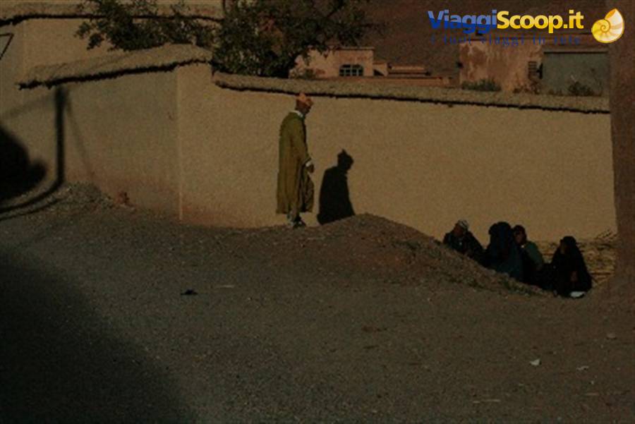 Man in green djellaba beside an earthen wall, women sitting in the shade