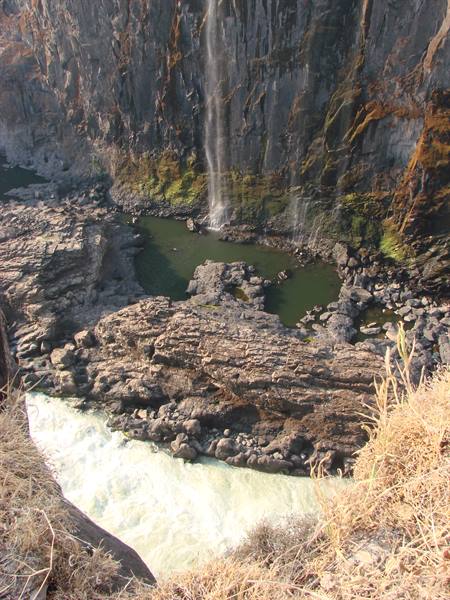 Thin waterfall on dark basalt cliff, rapids flowing over rocky riverbed