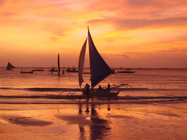 Sailboat with triangular sail on reflective beach, orange sky, fishermen pushing, Boracay