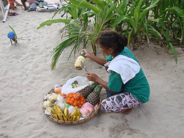 Woman crouching on sand cuts a pineapple, tray of fresh fruit