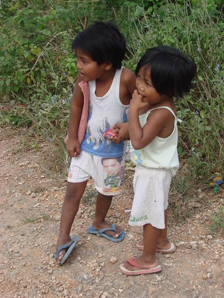 Two children in tank tops and flip-flops on a dirt path by bushes