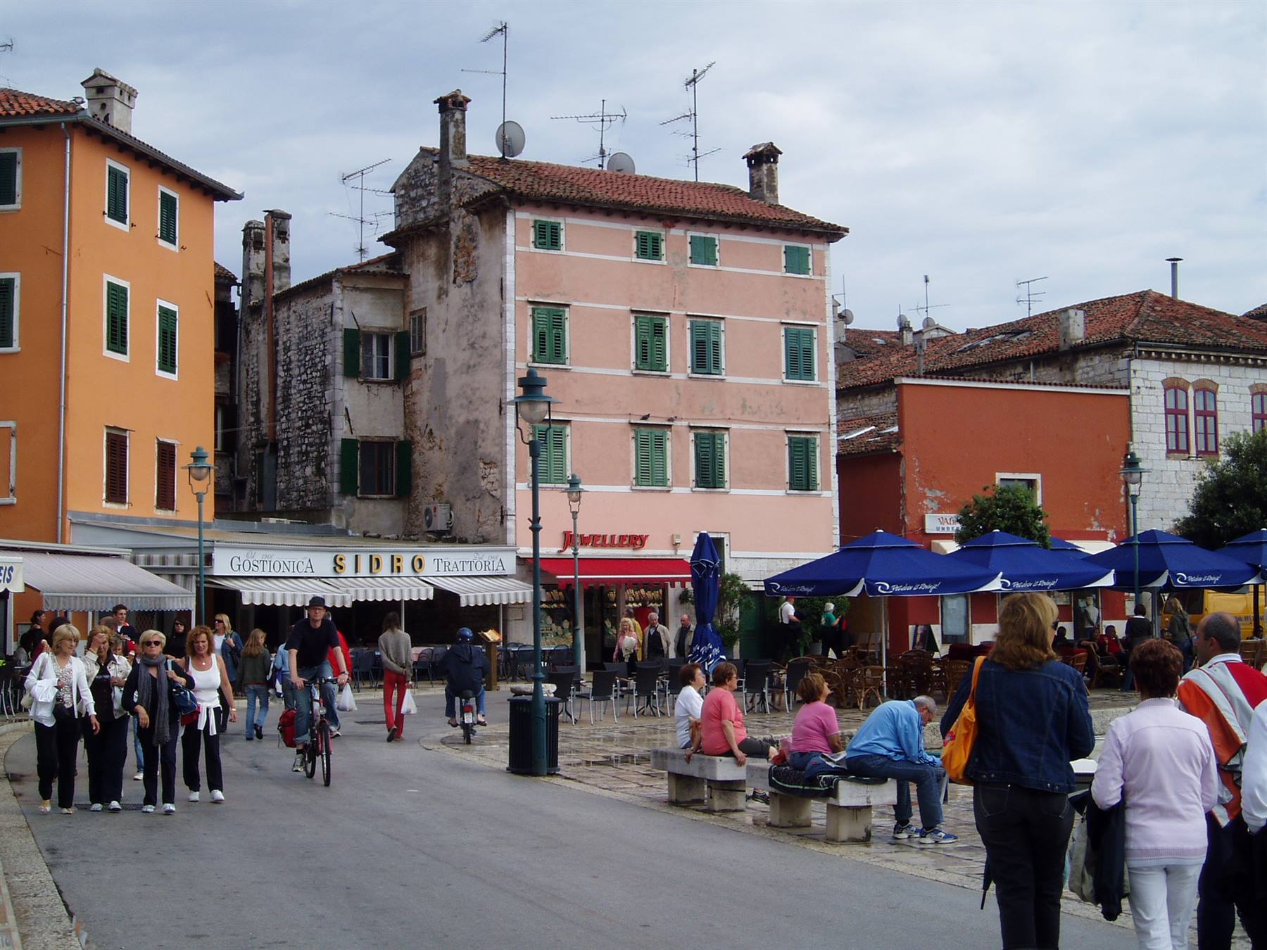 Tourists stroll a square with old houses, blue umbrellas and people sitting