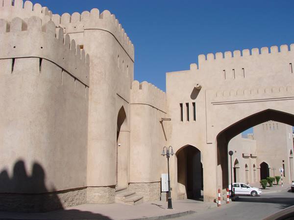 Mud-brick fort with crenellations and large arched gateway, cars passing beneath