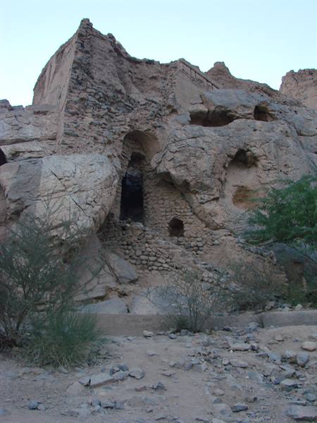 Ancient stone and mud ruins carved into rock, dry bushes below