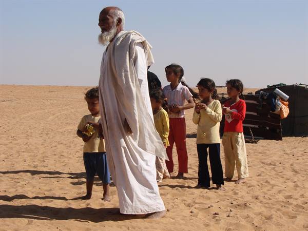 Elderly man in white robe with barefoot children on desert sand
