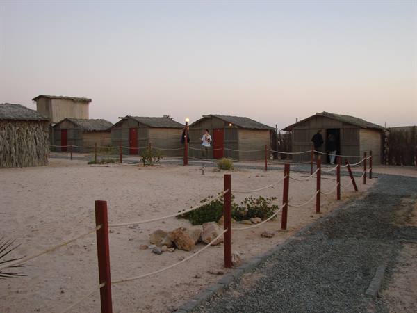 Wooden and straw huts on sand, tourists entering at dusk