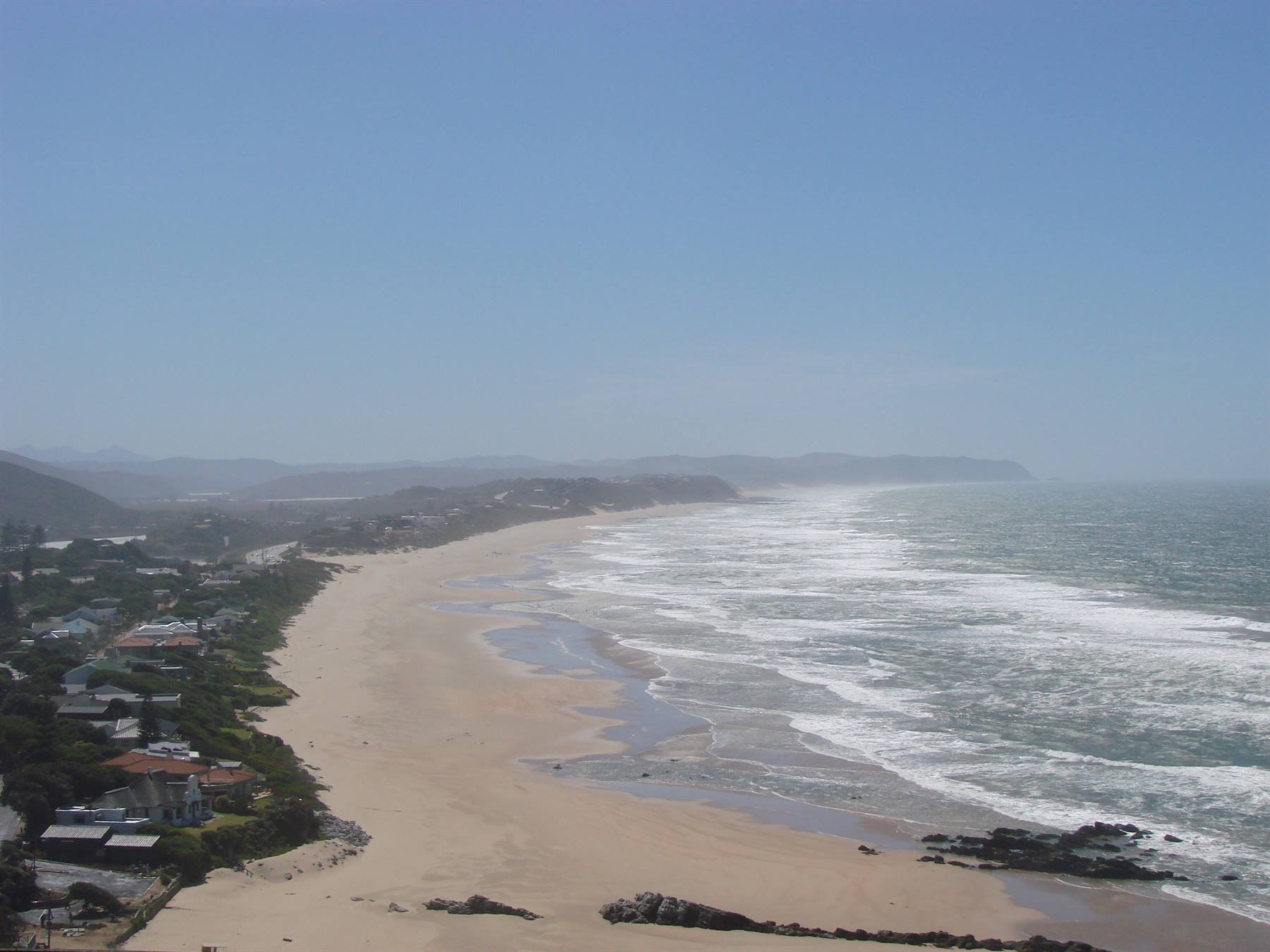 White waves on sandy beach beside houses among trees under blue sky