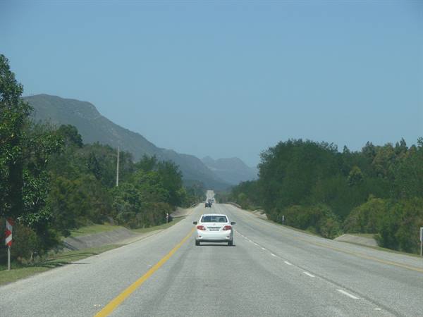 White car on asphalt road toward mountains, green trees, clear sky