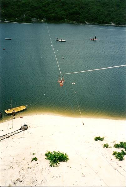 Person on a zipline over a green lake between sand dunes, boats waiting
