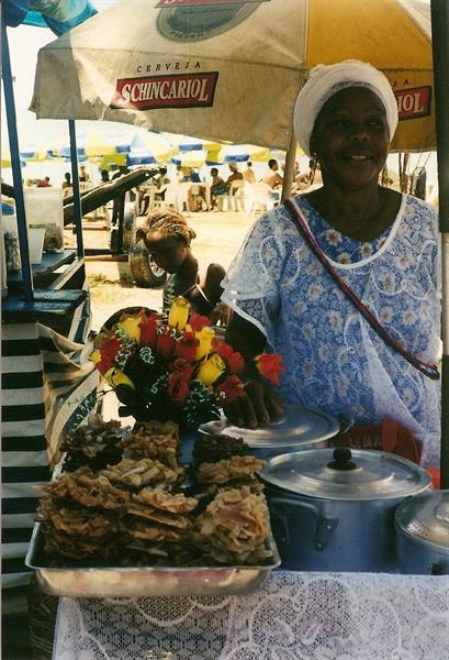 Smiling woman vendor in white and blue lace sells sweets under a Schincariol beach umbrella