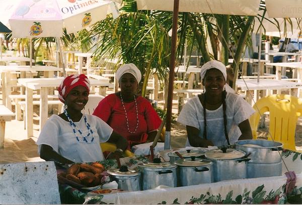 Women in headwraps sell food from aluminum pots under beach umbrellas