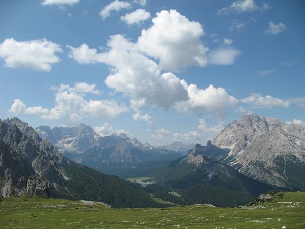 Green meadow and Dolomite peaks under white clouds, forested valley below