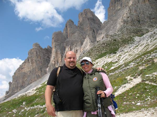 Smiling couple with hiking poles beneath jagged Dolomite peaks, blue cloudy sky