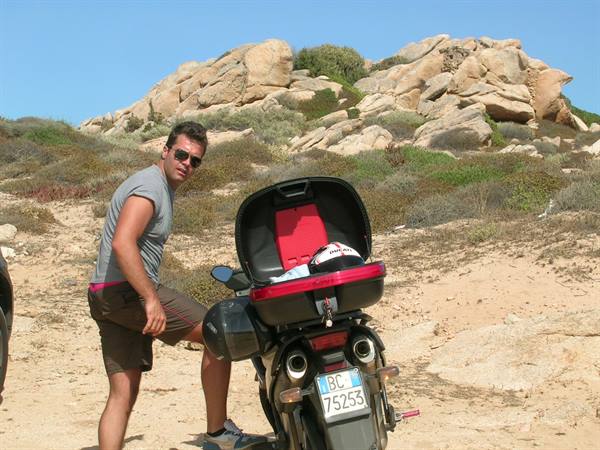 Man in sunglasses beside a motorcycle with open top case, granite rocks and Mediterranean scrub