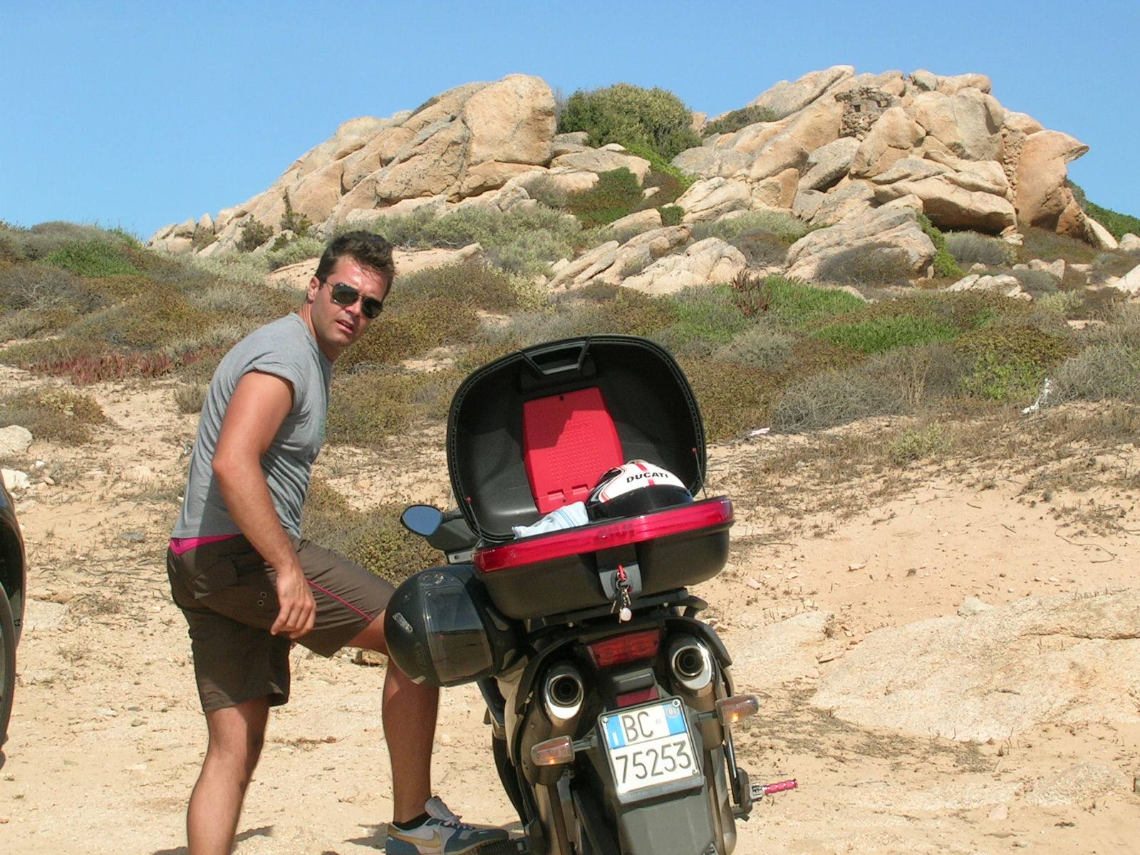 Man in sunglasses beside a motorcycle with open top case, granite rocks and Mediterranean scrub