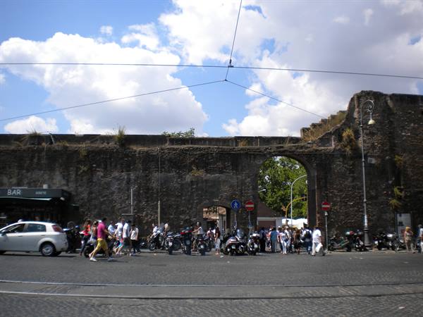 Pedestrians and scooters beneath an ancient brick gate in Rome